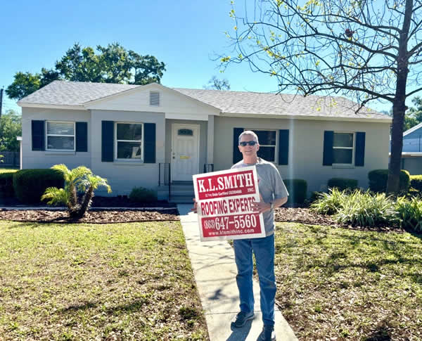 Satisfied client with a K.L. Smith Roofing Experts sign in front of a home with a newly replaced roof, symbolizing quality service and customer satisfaction.